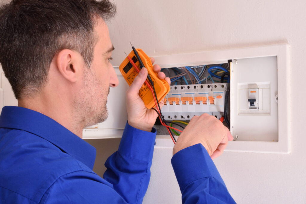 Professional electrician in blue uniform uses orange digital multimeter to test electrical panel with orange circuit breakers.