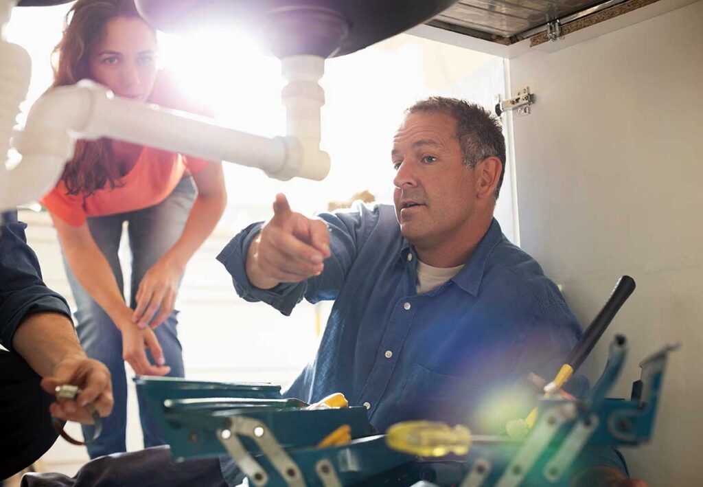 Plumbing technician instructing a group on holiday plumbing maintenance, pointing to explain repair techniques with tools visible on workbench.