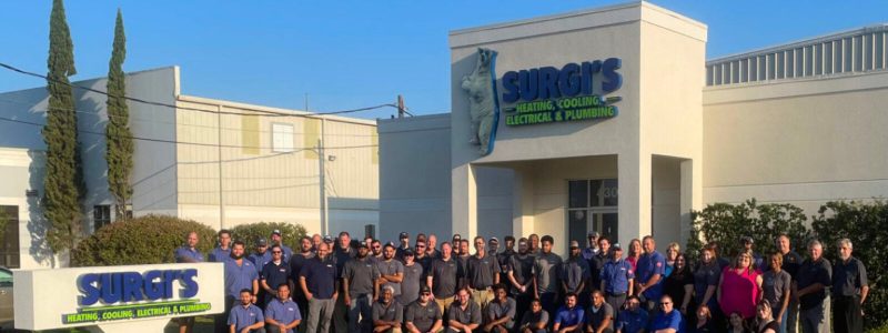 Large group of HVAC technicians and staff posed outside Surgi's Heating, Cooling, Electrical & Plumbing service building in New Orleans.